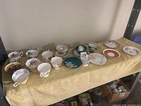 View of multiple orphan tea cups and saucers laid out on a yellow towel, showing a variety of patterns and colors.
