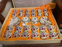 Wide shot of the tea set box showing the 12 teacups, 12 demitasses, 12 saucers, and 2 sugar bowls with spoons arranged in original packaging with orange fabric lining.