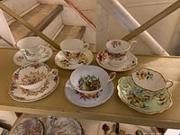 Six assorted floral-patterned bone china teacups with matching saucers placed on a shelf, showcasing varying floral designs and gold trim edges.