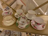 Overall display of six vintage porcelain teacups and saucers on shelf showing floral patterns and gold accents.