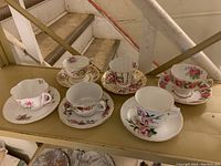 Six assorted vintage teacups and saucers arranged on wooden steps showing floral patterns and gold trim.