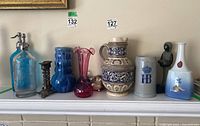 Full view of all 11 decorative items arranged on a mantle showing ceramic pitcher, two vintage seltzer bottles, Royal Copenhagen vase, glassware, candlesticks, stone carving