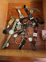 View of drawer showing about 16 assorted men's and women's wristwatches and one pocket watch. Various metal and leather bands in colors such as black, brown, green, and pink. Mix of dial styles, including digital and analog.