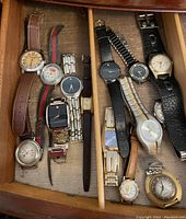 Top down view of 13 assorted men's and women's watches resting in a drawer, showing various brands, dial faces, and strap materials.