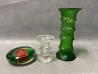 Three vintage glass items displayed on neutral background: oval paperweight with red flower, clear ribbed glass candleholder, and emerald green crinkle glass vase.