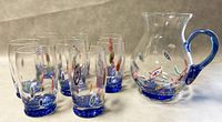 Glassware set of one clear Murano pitcher and eight tumblers on a neutral background. Tumblers and pitcher have blue bases and colorful millefiori glass patterns.