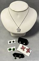 13 pieces of jewelry displayed on white necklace bust and flat surface, including necklace with butterfly pendant, several pairs of earrings in green faux stone, pearl, pink crystal by CORO.