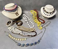 Wide view of vintage costume jewelry lot showing necklaces in various colors and two bangles, displayed on necklace stands and laid on table.
