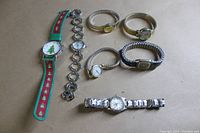 Photo showing all seven watches laying flat on a surface, displaying a variety of bands including fabric, expansion, and link styles in silver and gold tones. The Christmas tree watch is clearly visible.