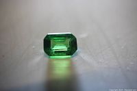 Close up photo of a loose emerald cut green garnet gemstone placed on a surface, showing its rectangular stepped facets and deep green color.