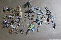 Overview of assorted costume jewelry items spread on a flat surface including earrings, pins, bracelets, charms, and rings in various styles and colors.
