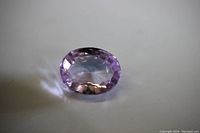 Oval cut pink sapphire loose gem lying flat on a plain white surface showing light pinkish-lilac color and transparency.