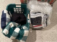 Photo shows a green plastic laundry basket containing two checkered blankets (green/white and blue/white) and a black fabric item, next to a packaged white down duvet with a red, white, and blue duvet cover.