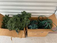 Two cardboard boxes containing green artificial pine garlands and green wire string lights with small bulbs, packed and ready for storage or use