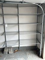Wide shot of one metal shelving unit mounted to wall with 5 adjustable wire mesh shelves. Concrete blocks on floor beneath shelving.