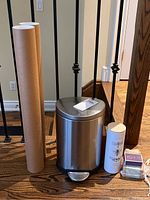 Wide view of stainless steel garbage can with pedal next to two tall brown cardboard tubes and candle tube.