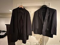 Front view of both jackets hanging side by side on a white door, showing the full length of the Bugatti black overcoat and the shorter Woolwich suit jacket with subtle checkered pattern.