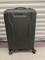 Front view of grey Samsonite hard case suitcase standing upright on four spinner wheels, showing brand name plaque, structural design on shell, and carry handle on top.