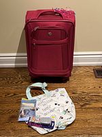 Front view of bright pink carry-on suitcase with front pockets and white tote bag with travel prints placed in front on wooden floor.