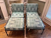 Pair of wrought iron chaise lounges with blue-grey checkered cushions placed indoors on hardwood floor.