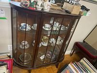 Front view of wood display cabinet showing curved glass doors with metal decoration and interior glass shelves containing various china teacups and saucers.