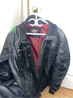 Front view of the black leather jacket hanging on a white hanger showing the maroon quilted lining and zipper details