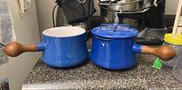 Two blue Dansk enamel pots with wooden handles placed side by side on a kitchen counter, one with a lid and one without, showing overall condition and size.