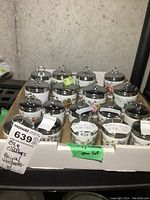 Overview of 18 Royal Worcester egg coddlers with silver lids, displayed in a cardboard tray, showing floral patterns including Lavinia and Bournemouth.