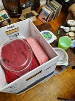 Open box containing a pink-daisy pattern rectangular casserole, glass lids and red packing cloth