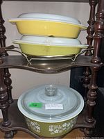 Two rectangular covered casseroles (one yellow with green leaf lid, one divided with sunflower lid) and one round floral bowl with glass knob lid arranged on shelf.