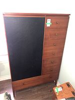 Front view of teak veneer armoire showing six drawers with brass knobs and a large black panel.
