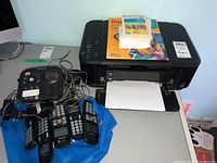 Canon PIXMA printer with four Panasonic cordless phones on a blue bag with wires and stacks of Kodak photo paper on top of the printer.