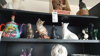 Shelf display showing assorted cat figurines and the colorful ceramic teapot with lid figurine on the top shelf.