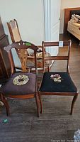Two vintage wooden chairs side by side, one carved backrest with embroidered dark seat, other ladder back with black fabric embroidered seat, both on wood floor.