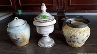 Three pieces of decorative china on dark surface: a scenic lidded jar, a covered urn, and a floral biscuit jar with metal lid.