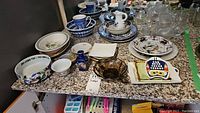 Photo showing various dishes, bowls, small pitchers and glassware of different shapes and patterns arranged on granite countertop.