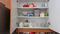Open cabinet shelves showing an assortment of kitchenware including bowls, glass ramekins, a Hen on Nest dish, and a napkin holder.