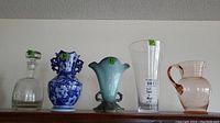 Five items arranged in a row on shelf: clear glass decanter, blue and white Hull vase, green Hull vase, tall clear glass vase, pink glass pitcher.