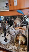 Five kettles including polished metal whistling kettles and electric coffee percolator with blue handle on countertop with original Luxbe packaging visible.