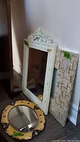 Three mirrors shown leaning against a wall; includes rectangular white frame mirror with floral crest, small oval framed mirror and rectangular textured frame mirror.