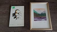 Two framed artworks lying next to each other on floor: indigenous print on left, landscape painting on right