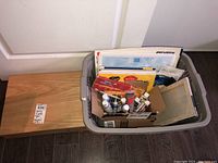 Wooden easel box closed next to a plastic container holding painting supplies including paint tubes, brushes, and canvas boards.