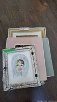 Stack of multiple framed paintings including one identified portrait in rustic frame