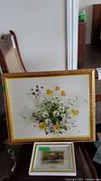 Two framed paintings stacked on a chair: larger floral painting on top and smaller painting on metal below.