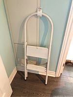 White metal step ladder standing upright against a door, showing full height and shape.