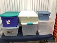 Photo showing seven assorted plastic storage bins stacked and placed on a black surface with a metal background. The bins are in different colors including purple, clear, blue, and white, varying in size.