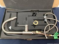 Top view of brushed finish kitchen faucet inside box, showing curved spout, detachable head, water lines, and mounting components.