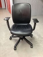 Front view of black leather office chair showing cushioned seat, backrest with lumbar contour, adjustable armrests, and wheeled black plastic base.