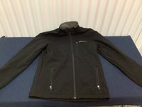 Full front view of black soft shell jacket laid flat showing zipper and pocket placement.