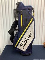 Front angled view showing the navy blue golf bag with white Titleist logo panel and yellow zipper detail, including attached tags.